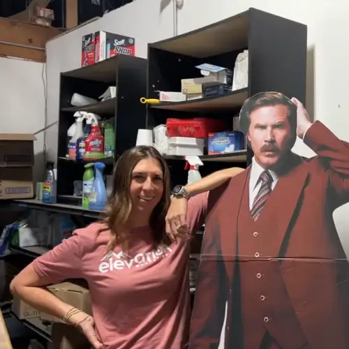 An Elevation Marketing team member smiling next to a Ron Burgundy cardboard cutout in an office supply area, showcasing the fun and creative atmosphere that fuels great Content Marketing.